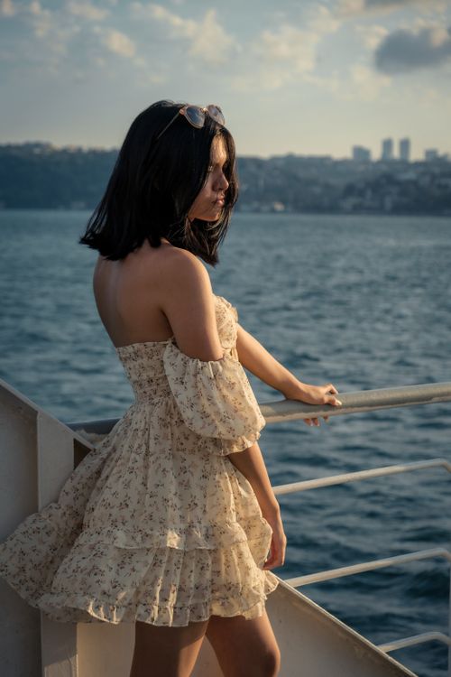 Free Young woman in an off-shoulder dress stands on a ferry, enjoying a scenic summer view. Stock Photo