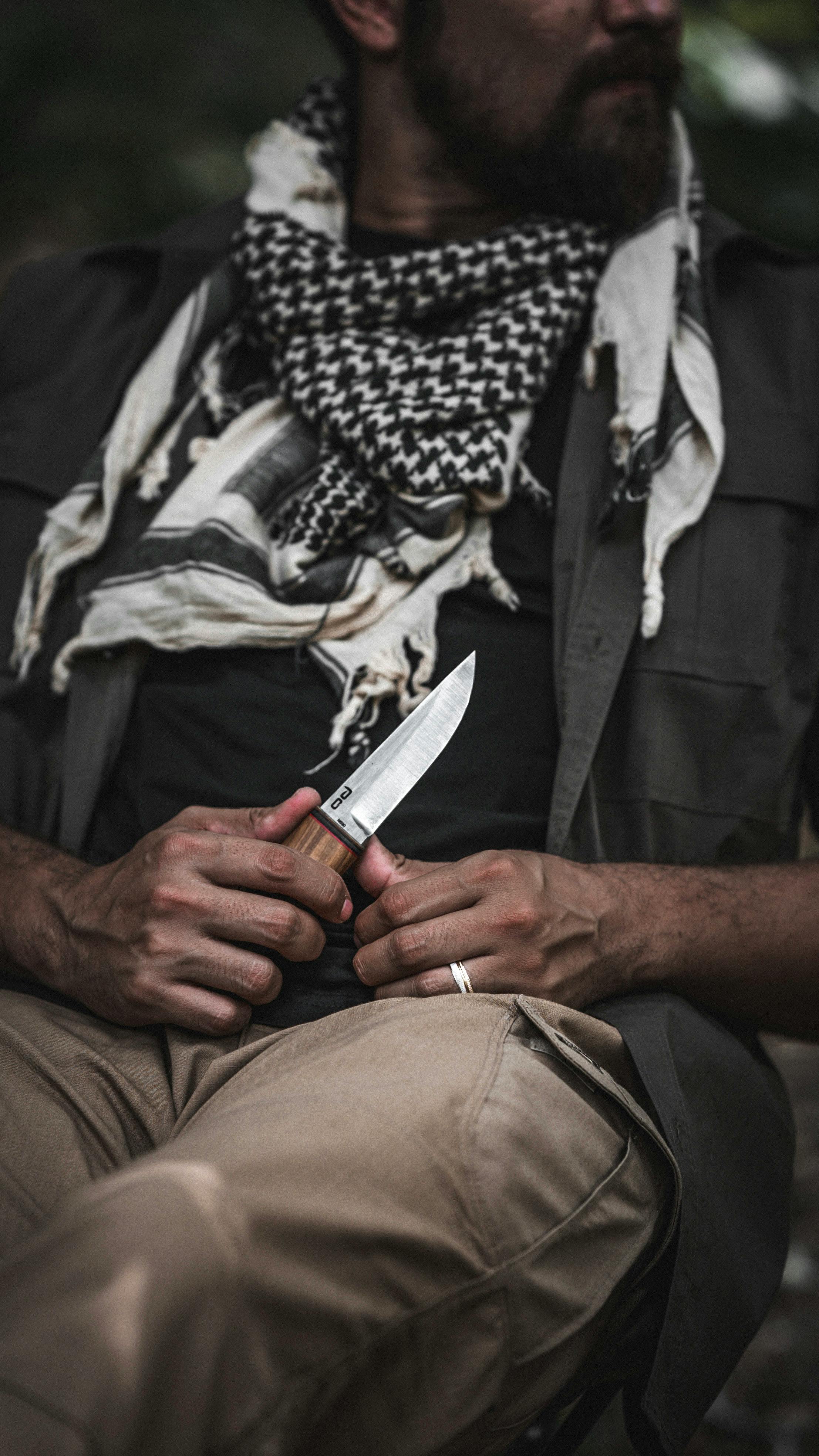 Man outdoors holding a knife, wearing a scarf, showcasing survival tools and gear.