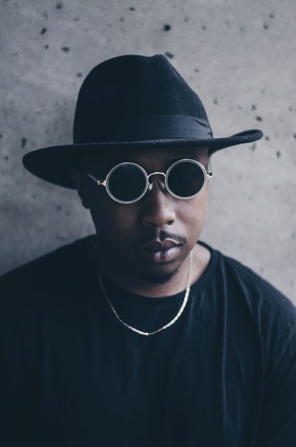 Fashionable portrait of a man wearing round sunglasses, hat, and necklace against a textured backdrop.