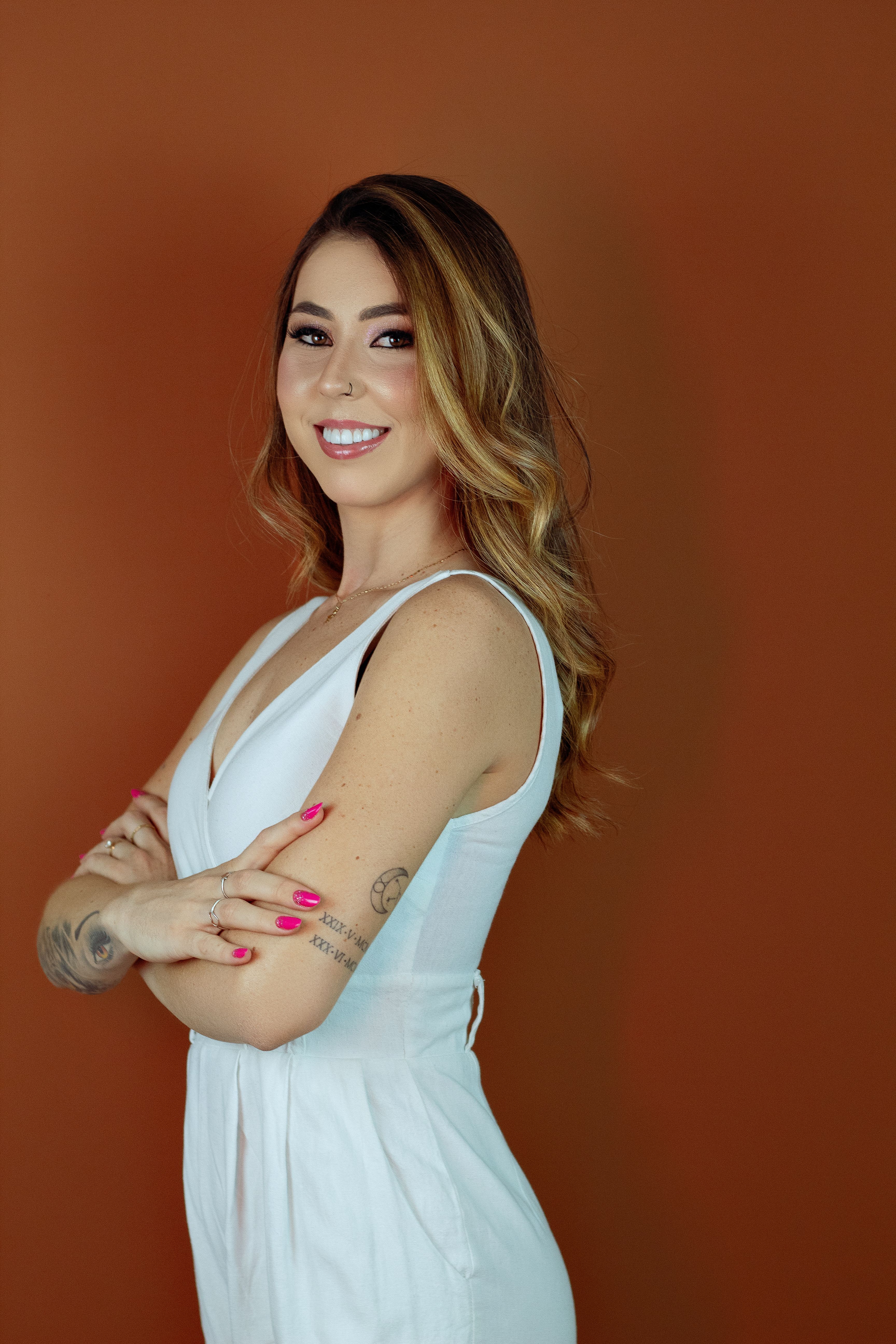 Portrait of a smiling woman posing confidently in a white dress against a brown backdrop.