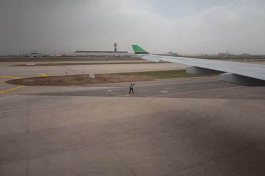 View of an airport runway with aircraft wing, control tower, and ground staff guiding on a cloudy day.