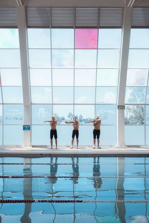 Free Three men in swimming trunks stretch by an indoor pool, preparing for training. Stock Photo
