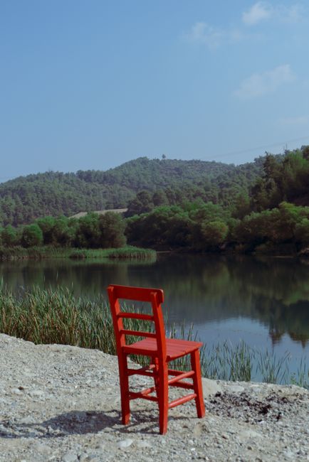 A lone red chair by a reflective lake, surrounded by forested hills in Aladağ, Türkiye.