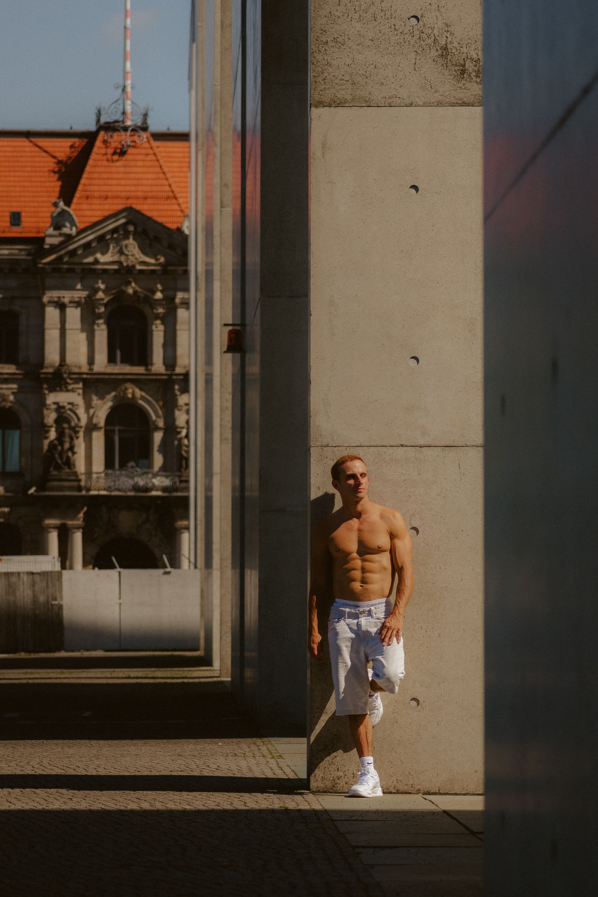 Athletic man posing shirtless next to modern and classic buildings.