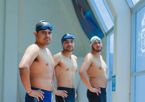 Free Three men stand in swimwear inside an indoor swimming pool area, ready for swimming activities. Stock Photo