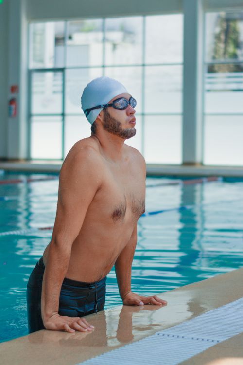 Free Focused male swimmer standing at the edge of an indoor swimming pool, wearing goggles and swim cap. Stock Photo