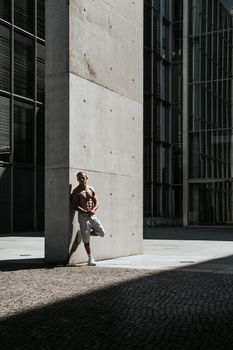 Shirtless muscular man posing by a concrete pillar in a modern urban area during daylight.