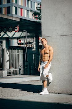 Muscular man posing shirtless in an urban environment, showcasing fitness and style.
