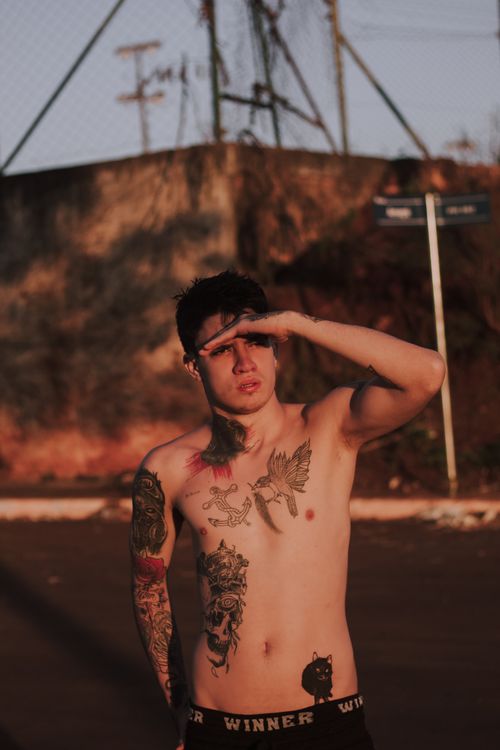 Free A tattooed man standing outdoors, shading his eyes in the sunlight, capturing a street vibe. Stock Photo