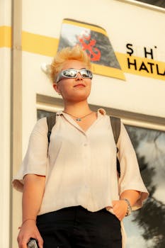 Casual portrait of a blonde woman wearing sunglasses, standing confidently in an urban setting during daytime.