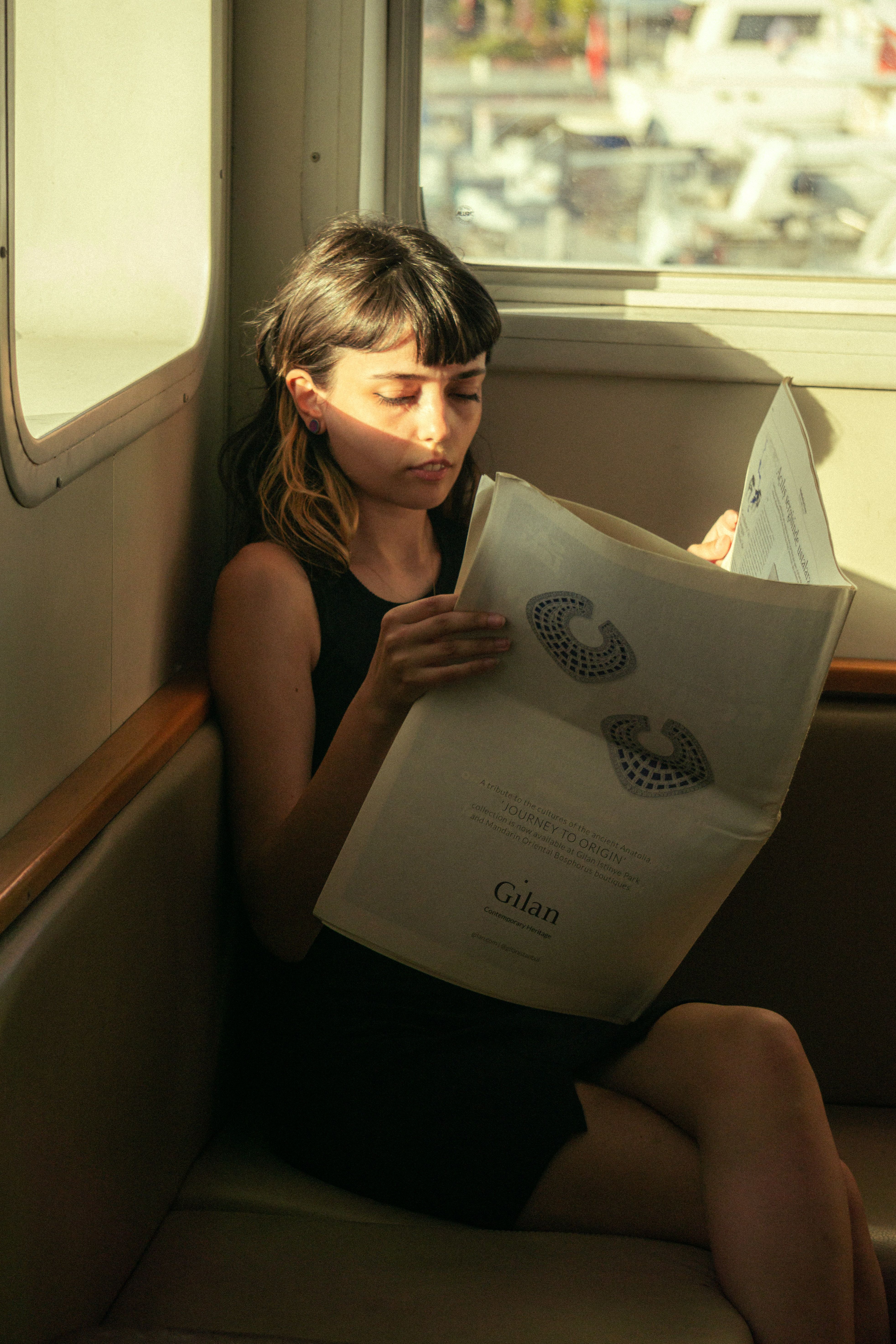 Young woman with brunette hair reading a newspaper in a sunlit train compartment, creating a calm and contemplative moment.