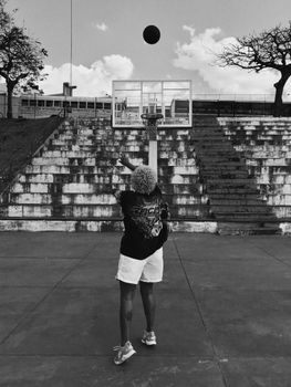 Adult shooting basketball outdoors in monochrome, showcasing athletic action and urban scenery.