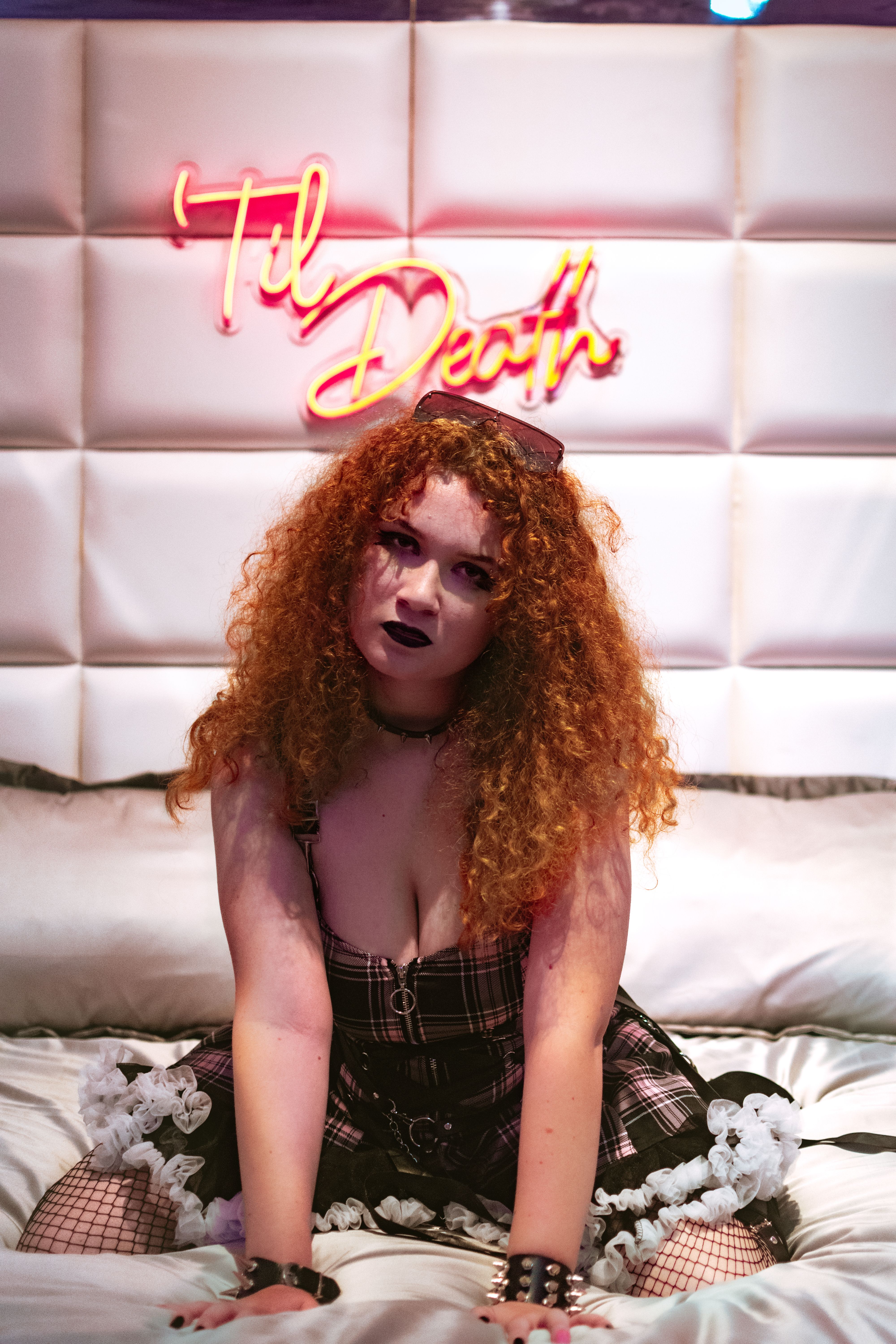 Redheaded woman in edgy attire sitting on bed with neon sign 'Til Death' in background.