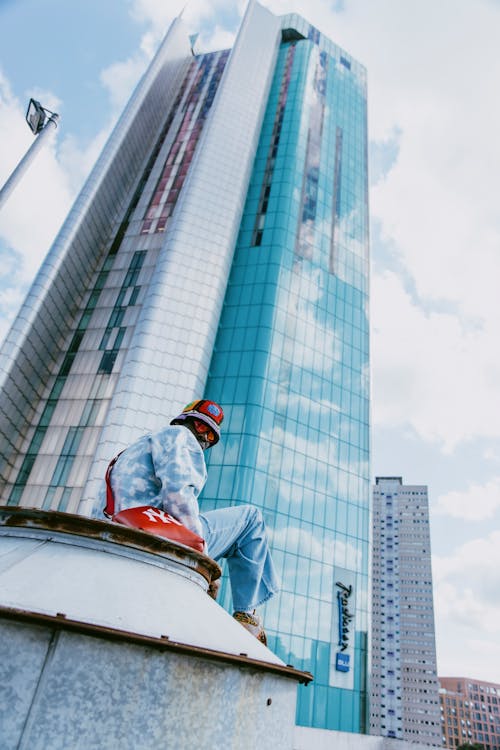 Young Man in Denim Shirt, Jeans, and Multicolored Bucket Hat Sitting near Skyscraper
