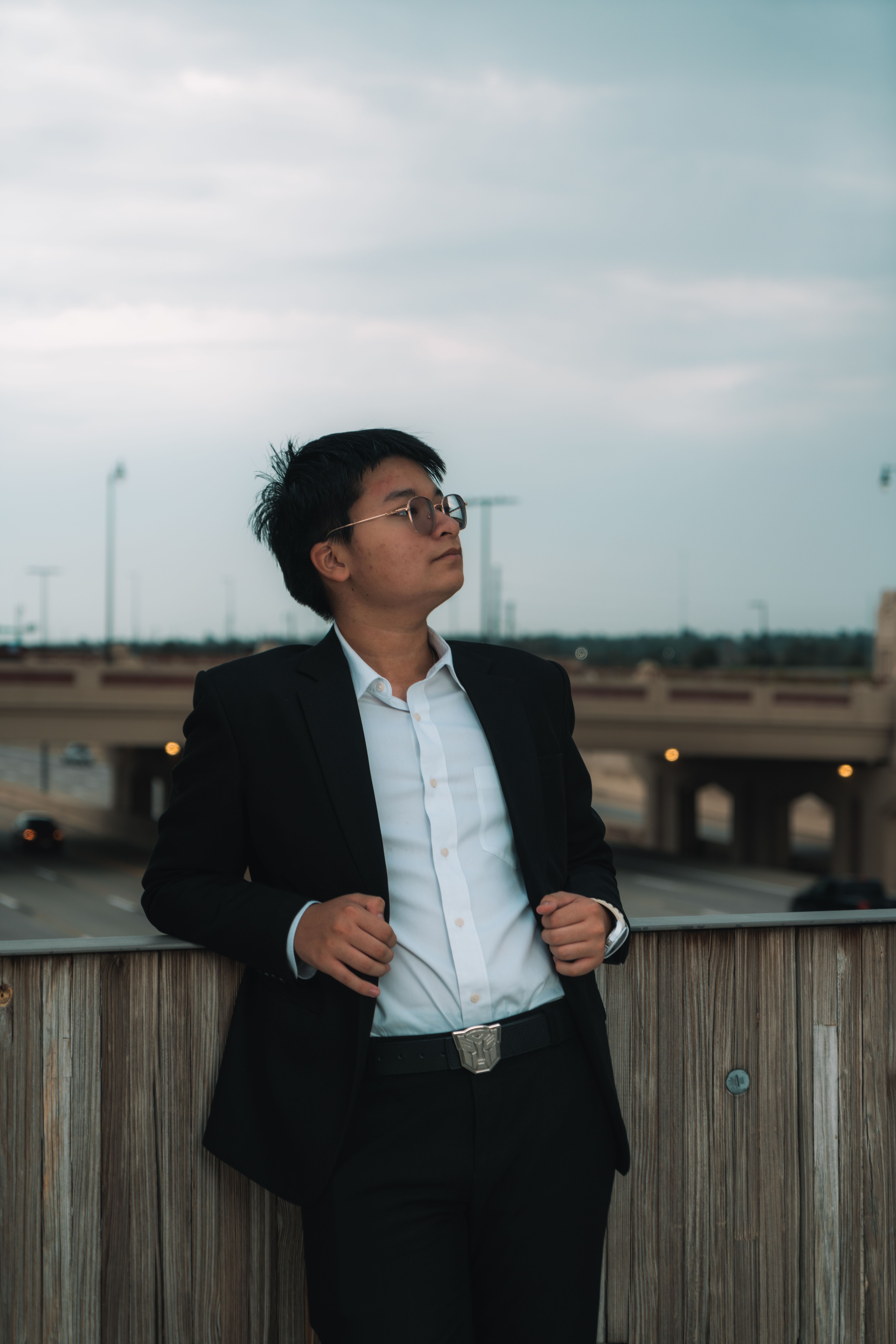 Asian man in a black suit poses stylishly against a balcony with an urban setting in the background.