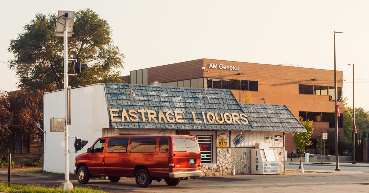 A vibrant street scene featuring a liquor store and parked vintage red van under a clear sky.