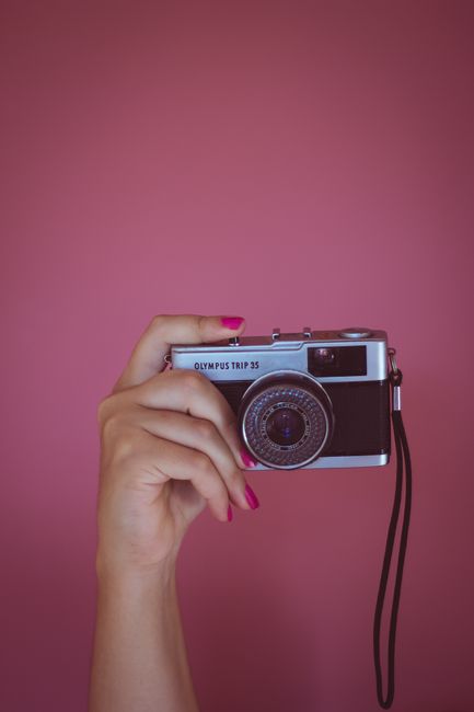 A retro Olympus Trip 35 camera held by a woman's hand against a pink backdrop.