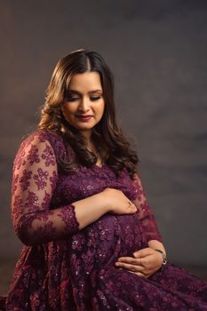 Beautiful studio portrait of a pregnant woman in a purple embroidered dress, embracing her belly. - Fotografer Sidoarjo