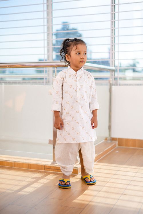 Free Young child wearing traditional clothing stands by a window with sunlight streaming through blinds. Stock Photo