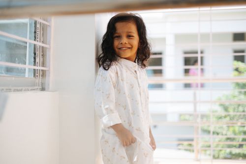 Free A smiling child in bright sunlight, dressed in white, standing indoors near a window. Stock Photo