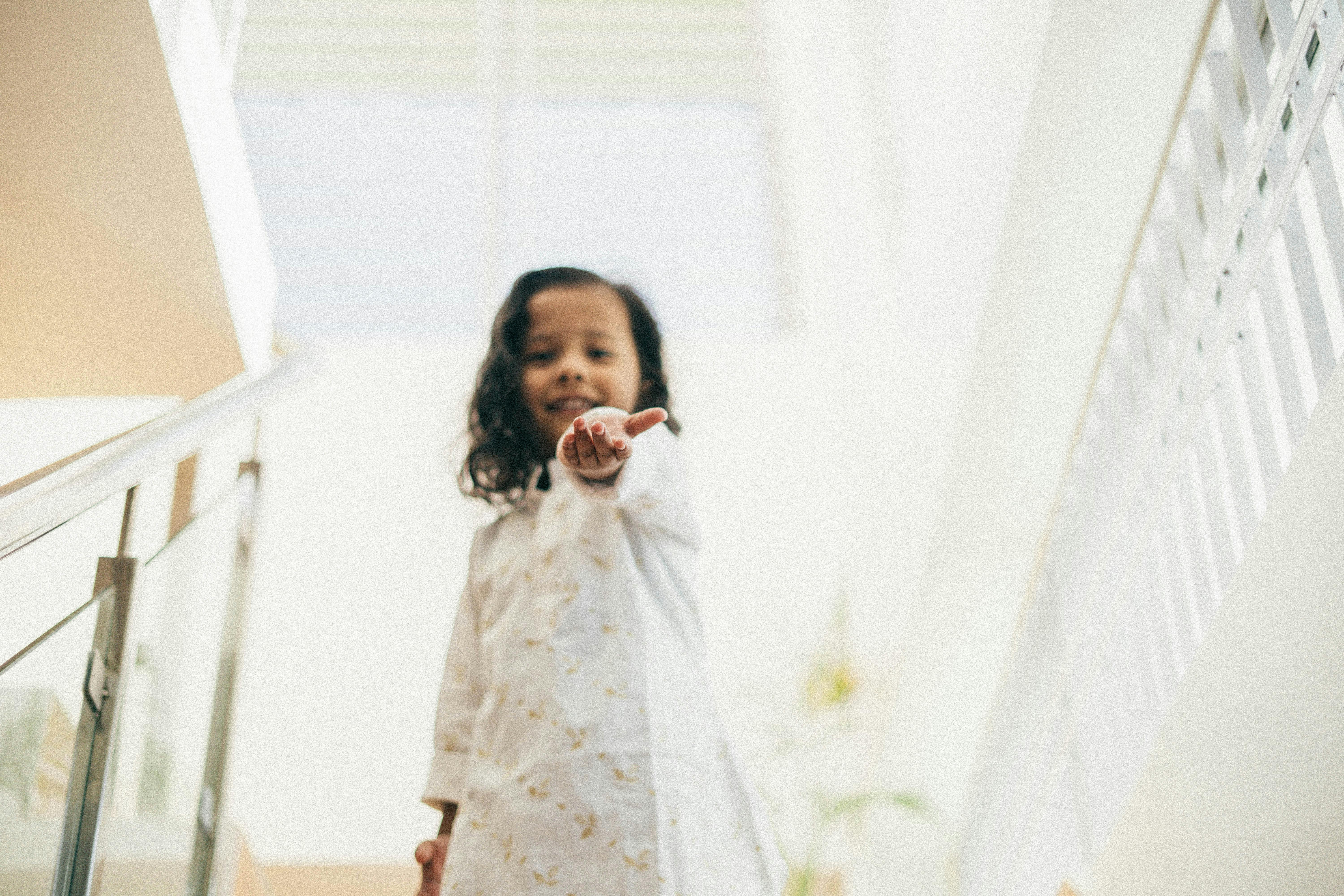 A young girl with a warm smile extends her hand while standing indoors on stairs.
