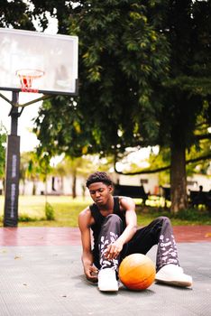 Young man sits on an outdoor basketball court tying his shoes, basketball nearby.
