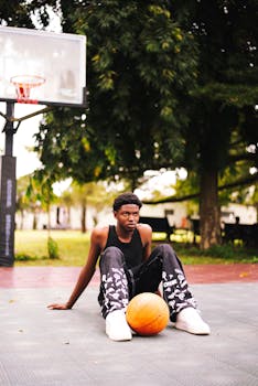 A young man sitting on a basketball court with a ball, surrounded by trees.