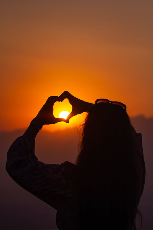 Silhouette of a Woman Making a Heart Shape with Her Hands on the Background of a Sunset Sky 