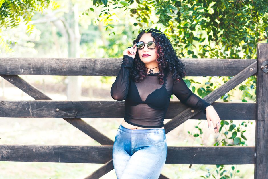 Confident woman with sunglasses poses by a wooden fence in a park setting.