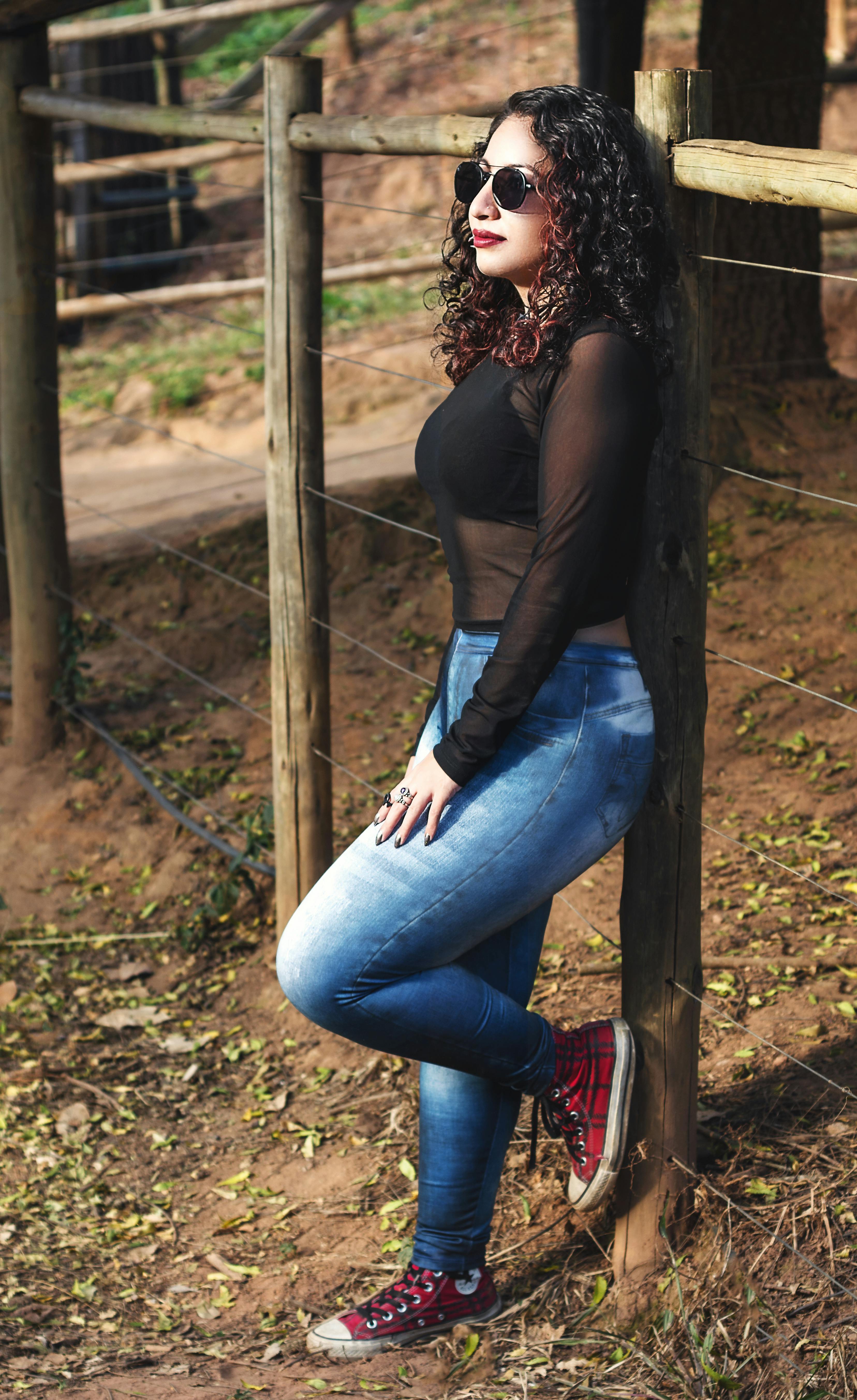 在一个阳光明媚的日子里，一位卷发、穿着时尚的年轻女子在木栅栏旁摆着姿势