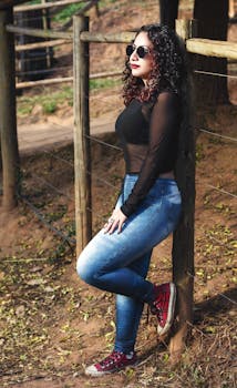 在一个阳光明媚的日子里，一位卷发、穿着时尚的年轻女子在木栅栏旁摆着姿势