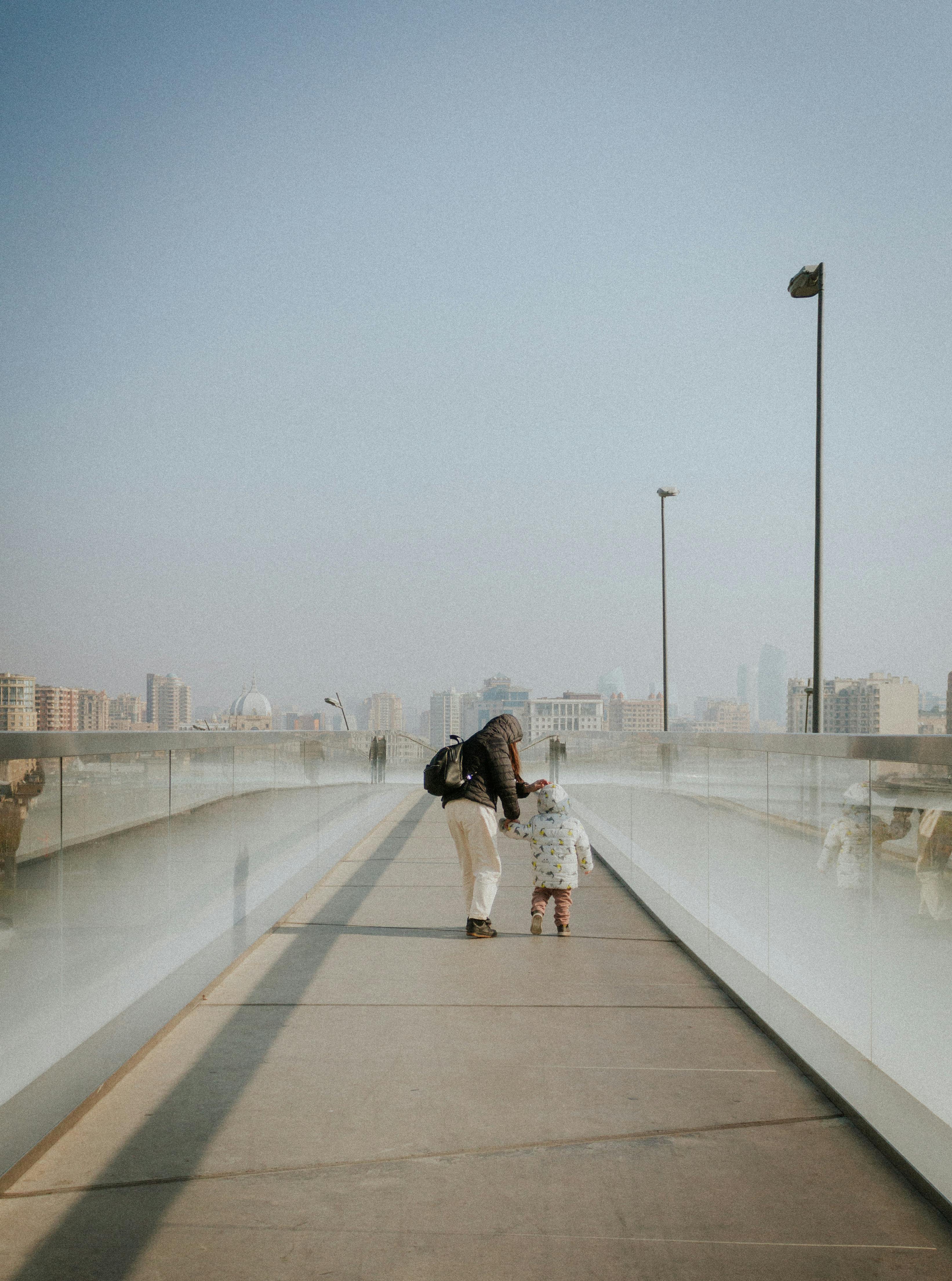 A woman and child walk hand in hand on a misty bridge towards a cityscape in the distance.