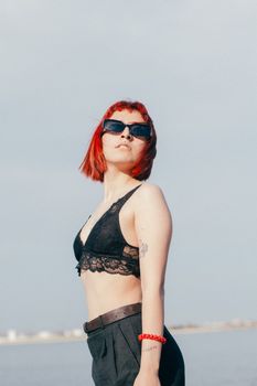 Fashionable young woman with red hair and sunglasses in a black bra posing outdoors.