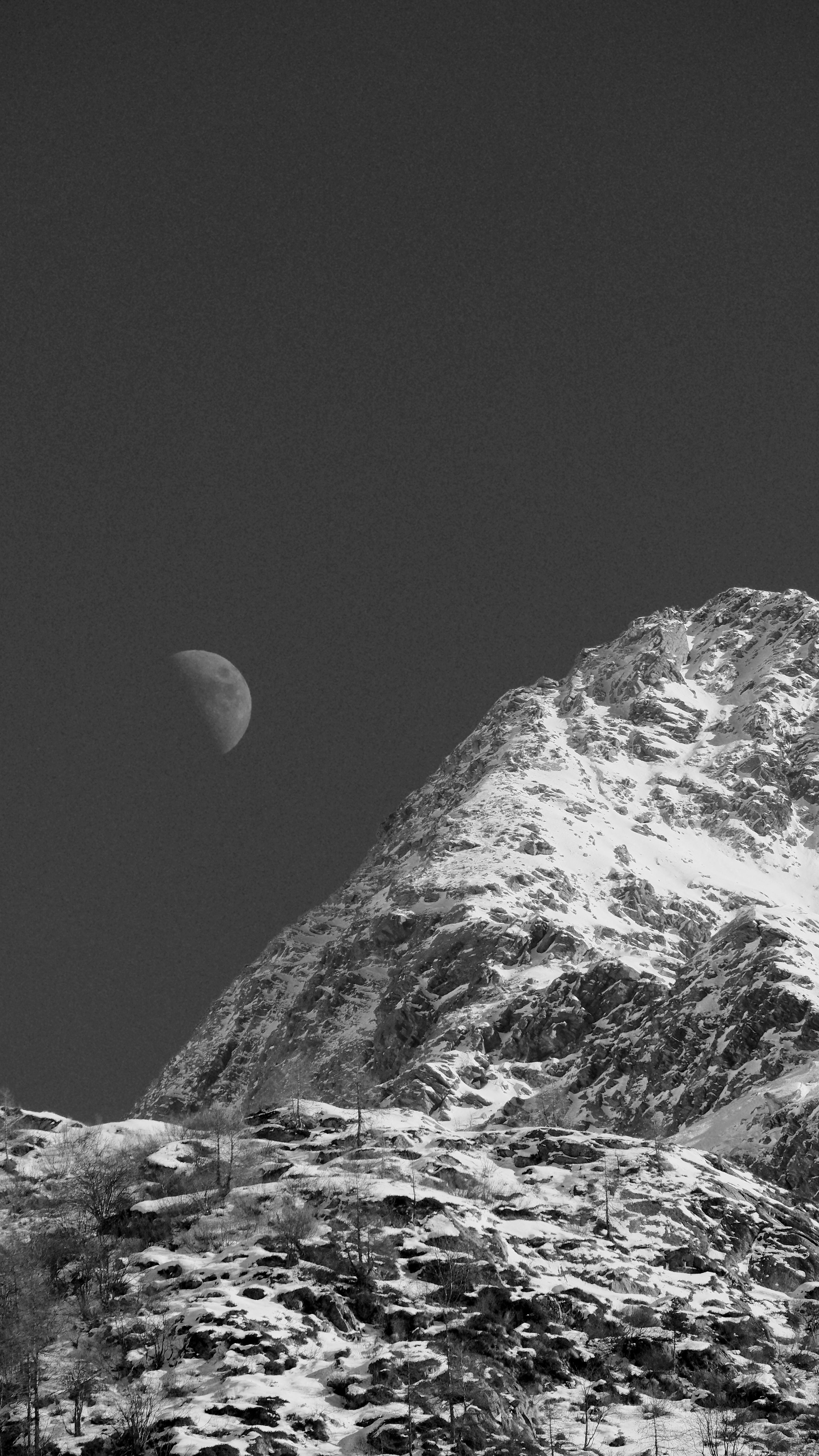 Black and white image of a snowy mountain with the moon in the background.