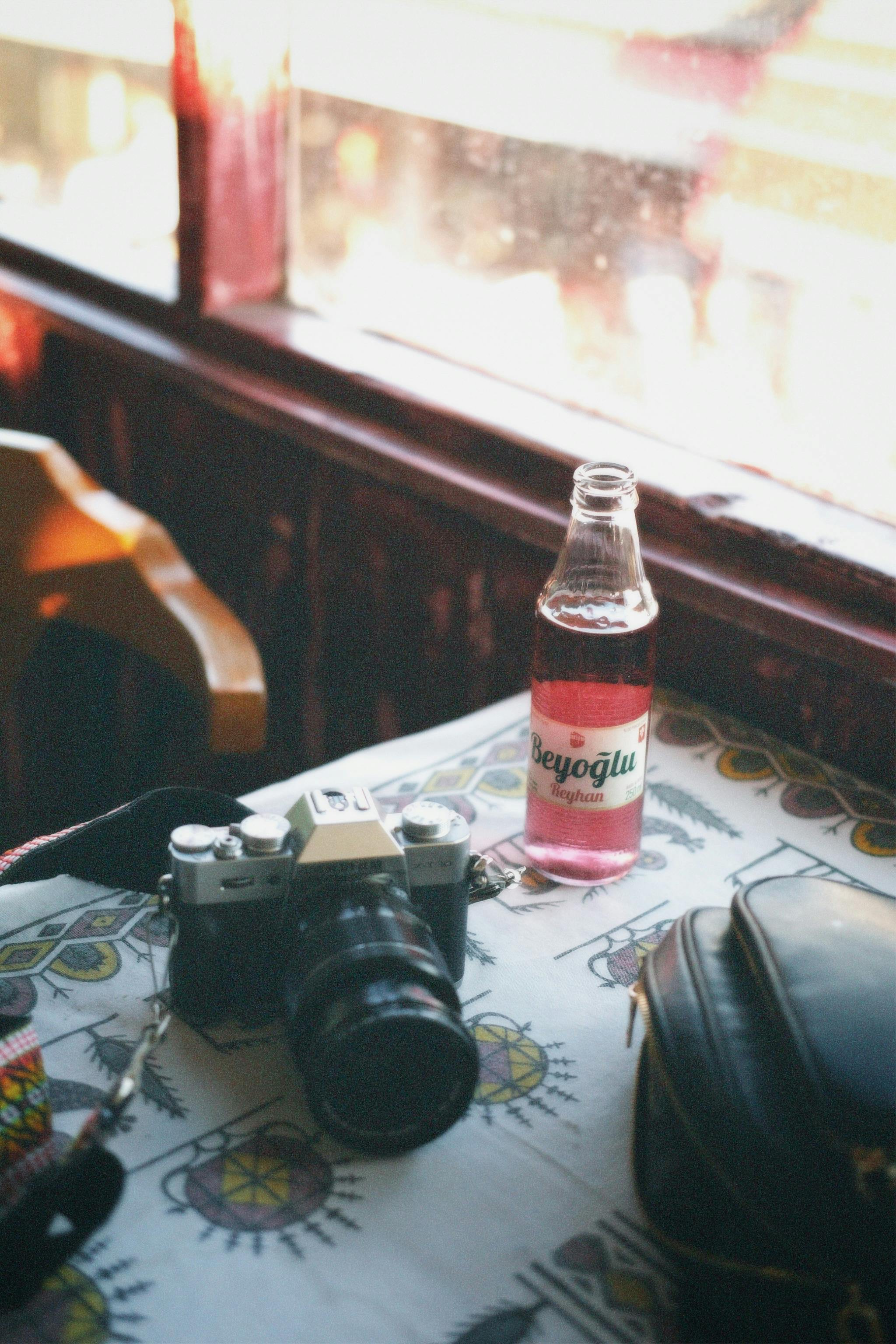 A vintage camera and Beyoglu drink on a patterned table beside a window in daylight.