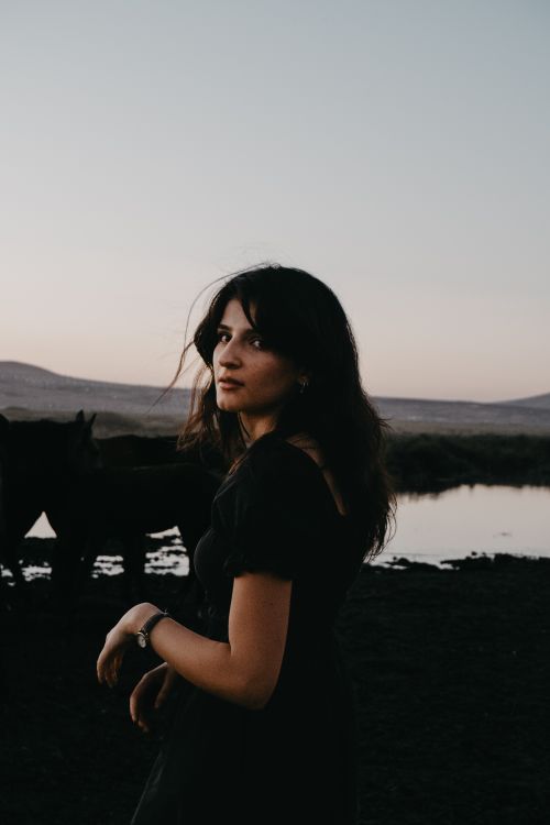 Free A beautiful woman stands gracefully by a river at sunset, capturing serene elegance. Stock Photo