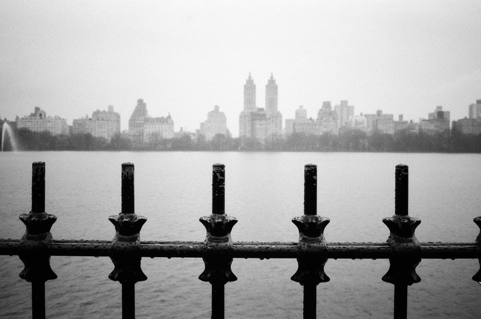 Black and white photo of NYC skyline and reservoir from behind a fence.