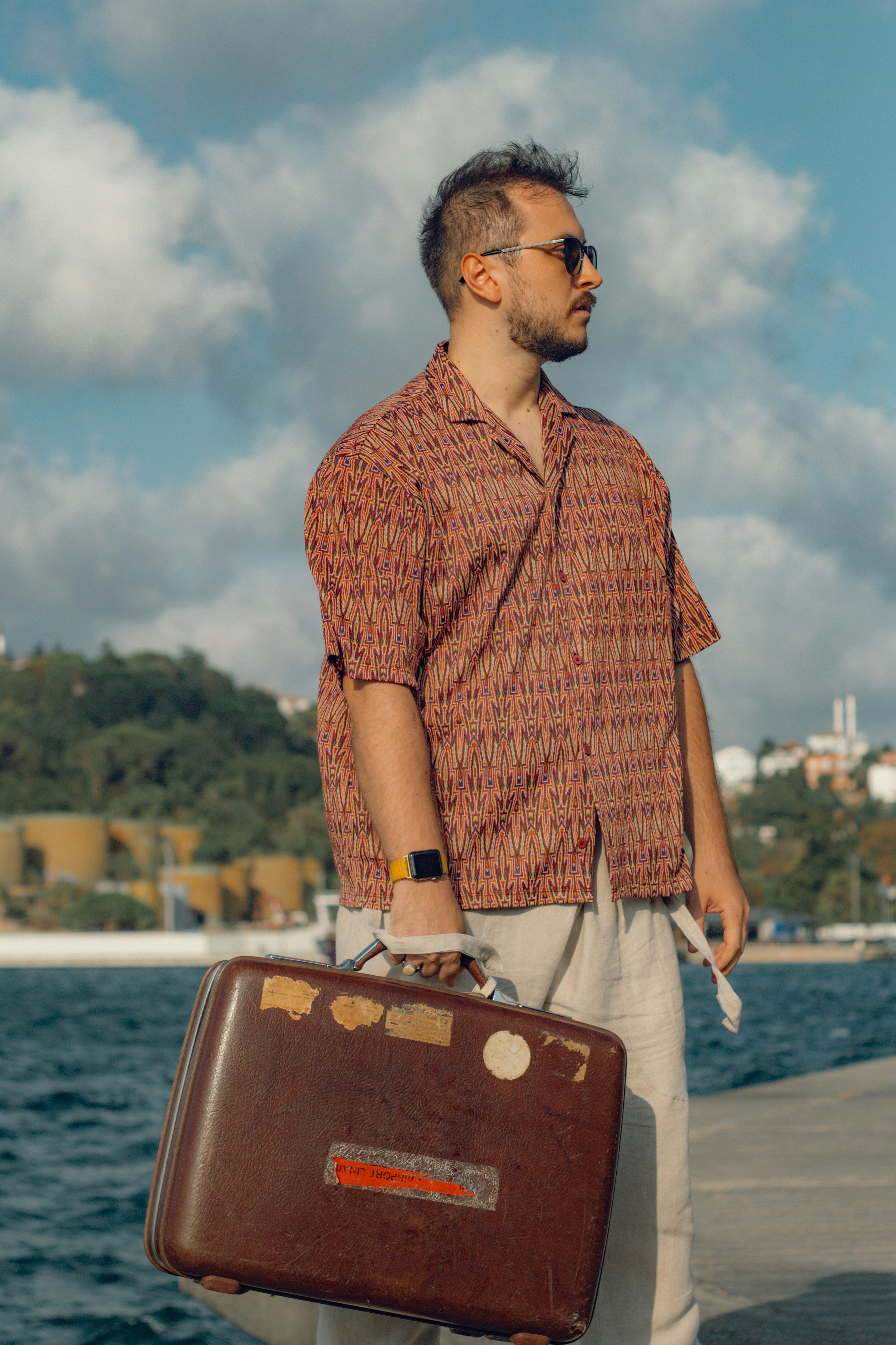 A young man stands by the İstanbul harbor with a suitcase, under bright summer sunlight.