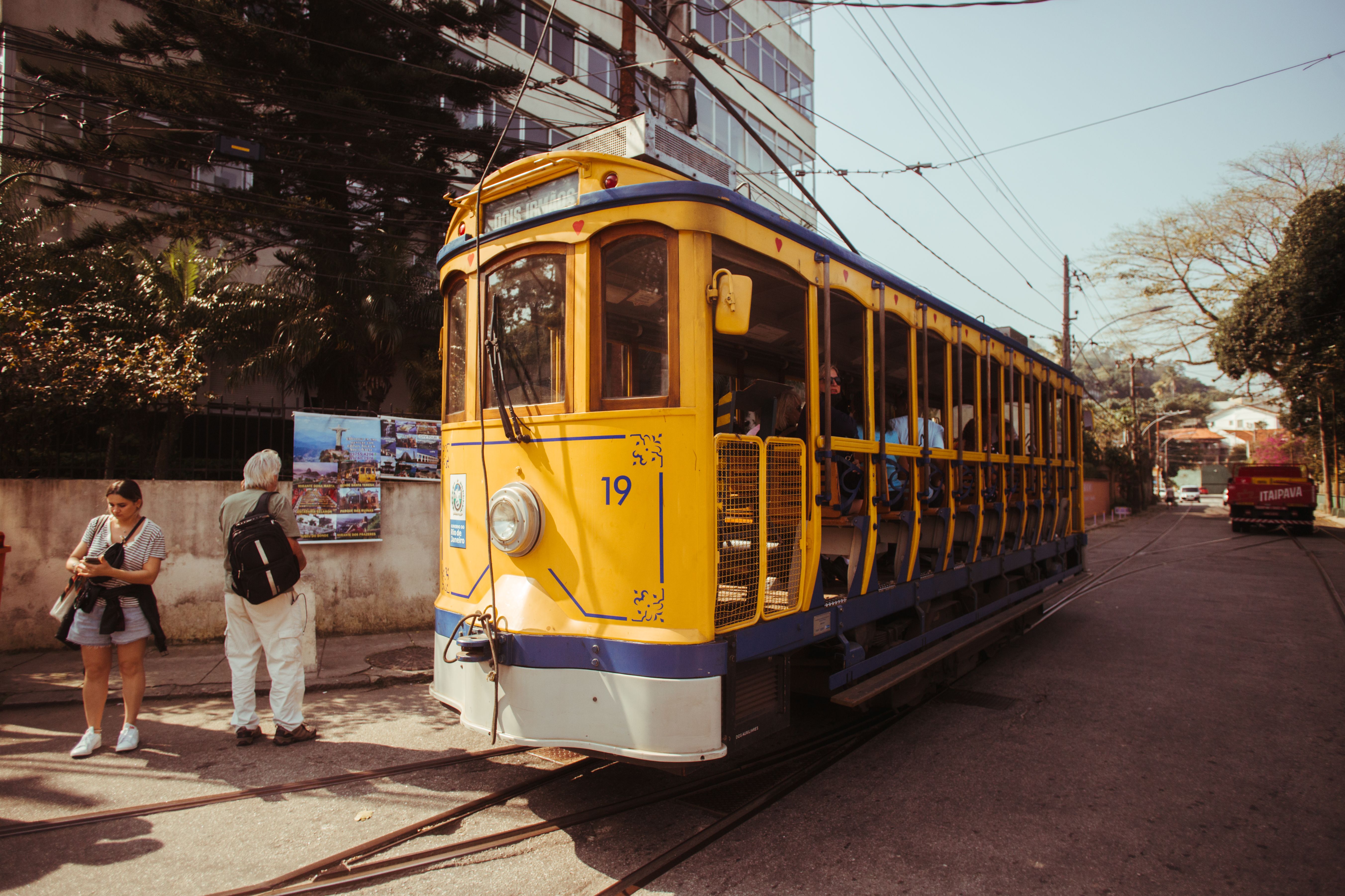 A yellow and blue open-sided tram numbered 19 beside a pavement, with two people standing by the track, overhead wires above and a red truck down the road