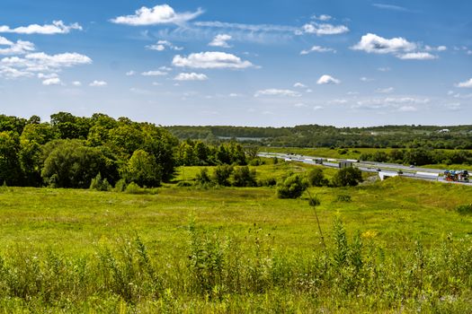 A wide view of a highway cutting through lush meadows and fields under a bright blue sky in Pickering, Canada.