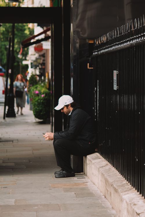 Free A man in a white cap sits on a sidewalk in London, engaged with his smartphone. Stock Photo
