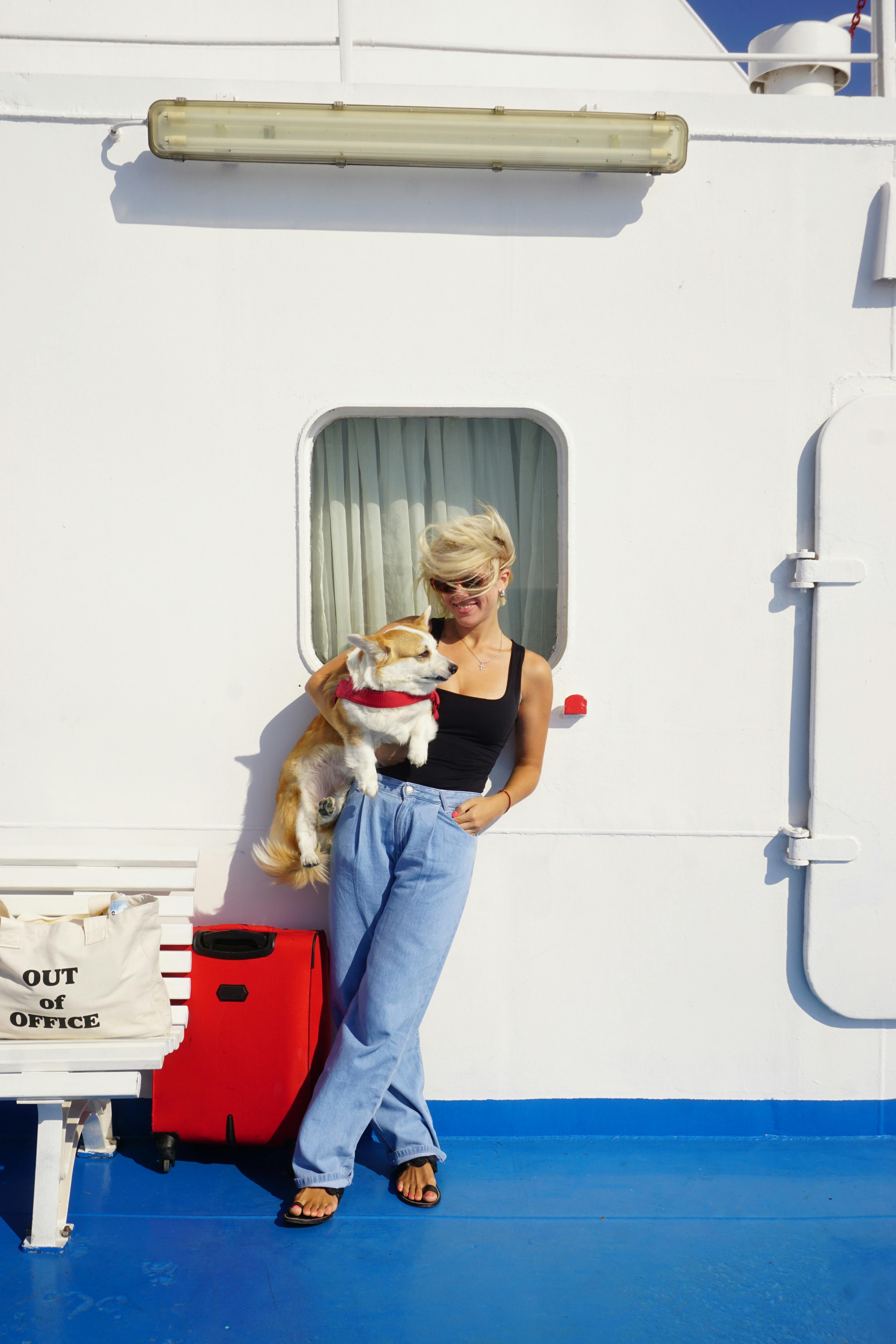 A woman holding a corgi dog on a ferry, with luggage and an 'Out of Office' tote bag.