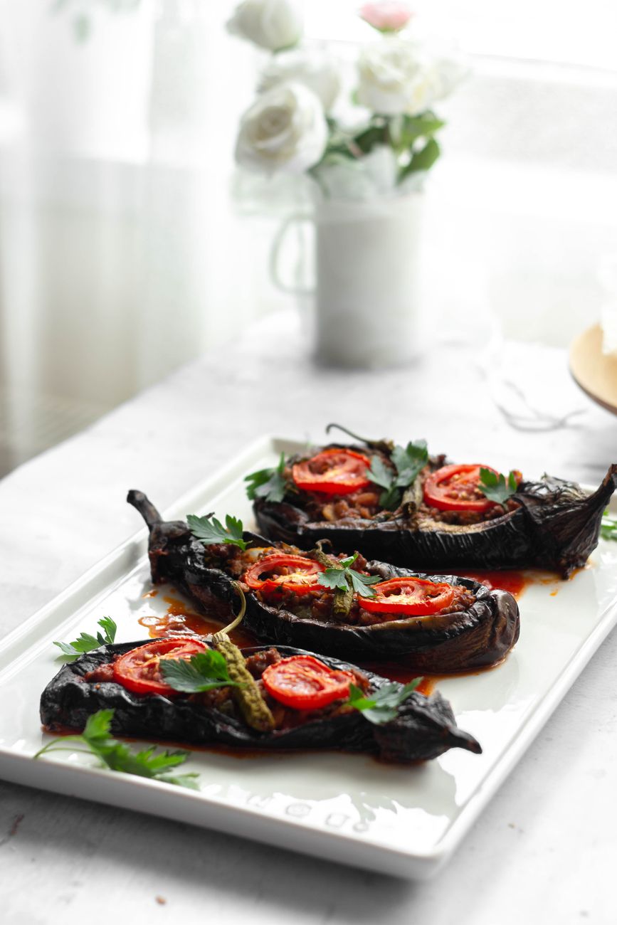 Four golden-brown roasted eggplant halves filled with spiced rice pilaf, garnished with fresh herbs and glistening with harissa oil on a rustic ceramic plate
