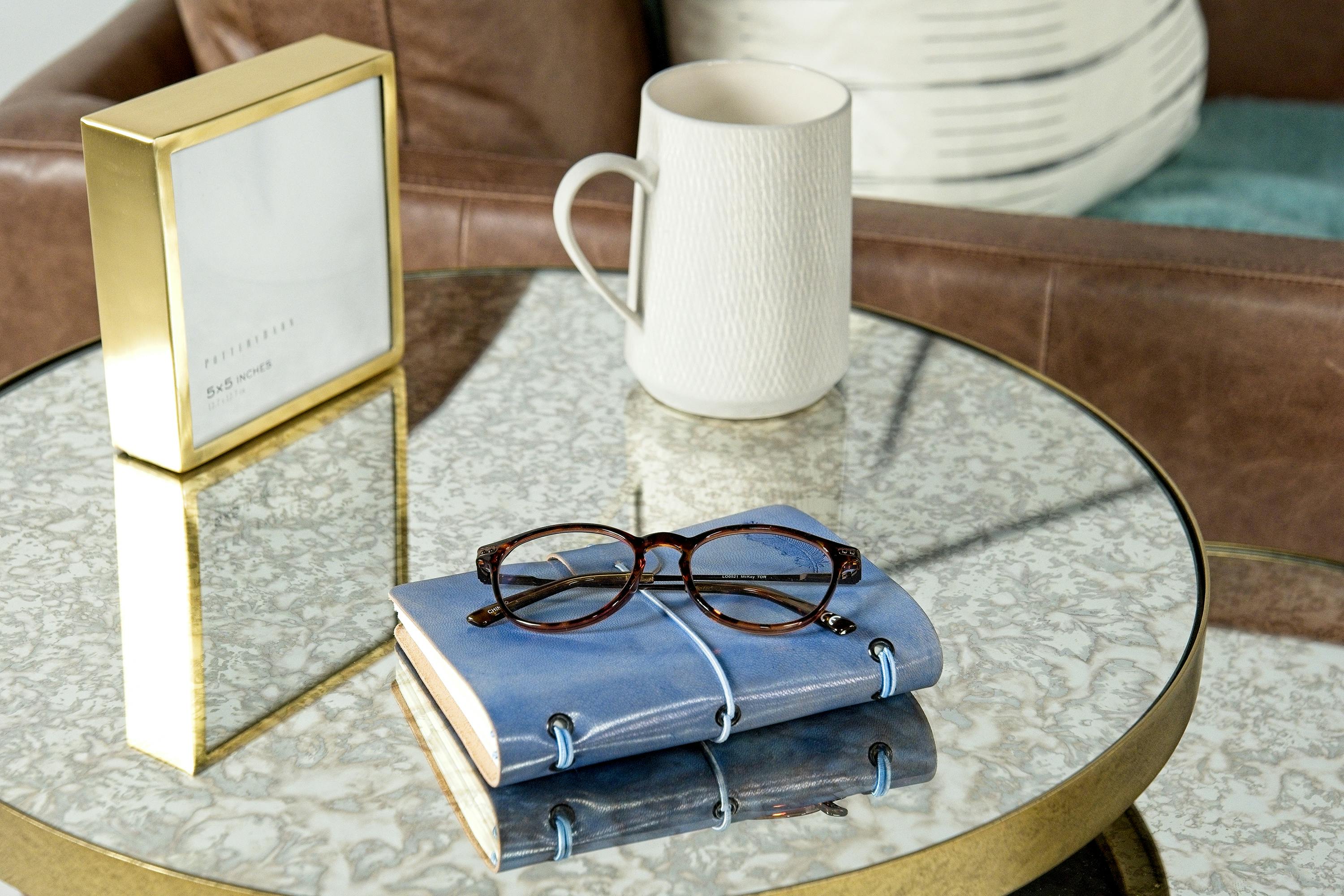 Cozy café setting with a mug, glasses, and notebook on a chic glass table.