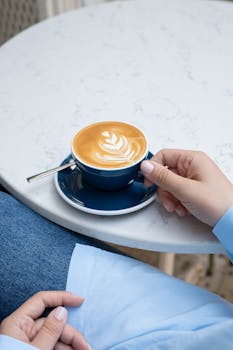 A stylish cappuccino with intricate latte art in a blue cup on a round table, held by a person in a blue shirt.
