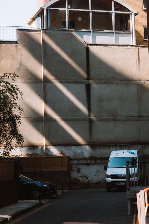Free Sunlit urban alley with vehicles, showcasing shadow patterns on a large wall. Stock Photo