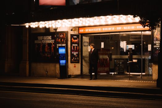A man waits outside a brightly lit theater entrance on a city street at night, capturing urban elegance.