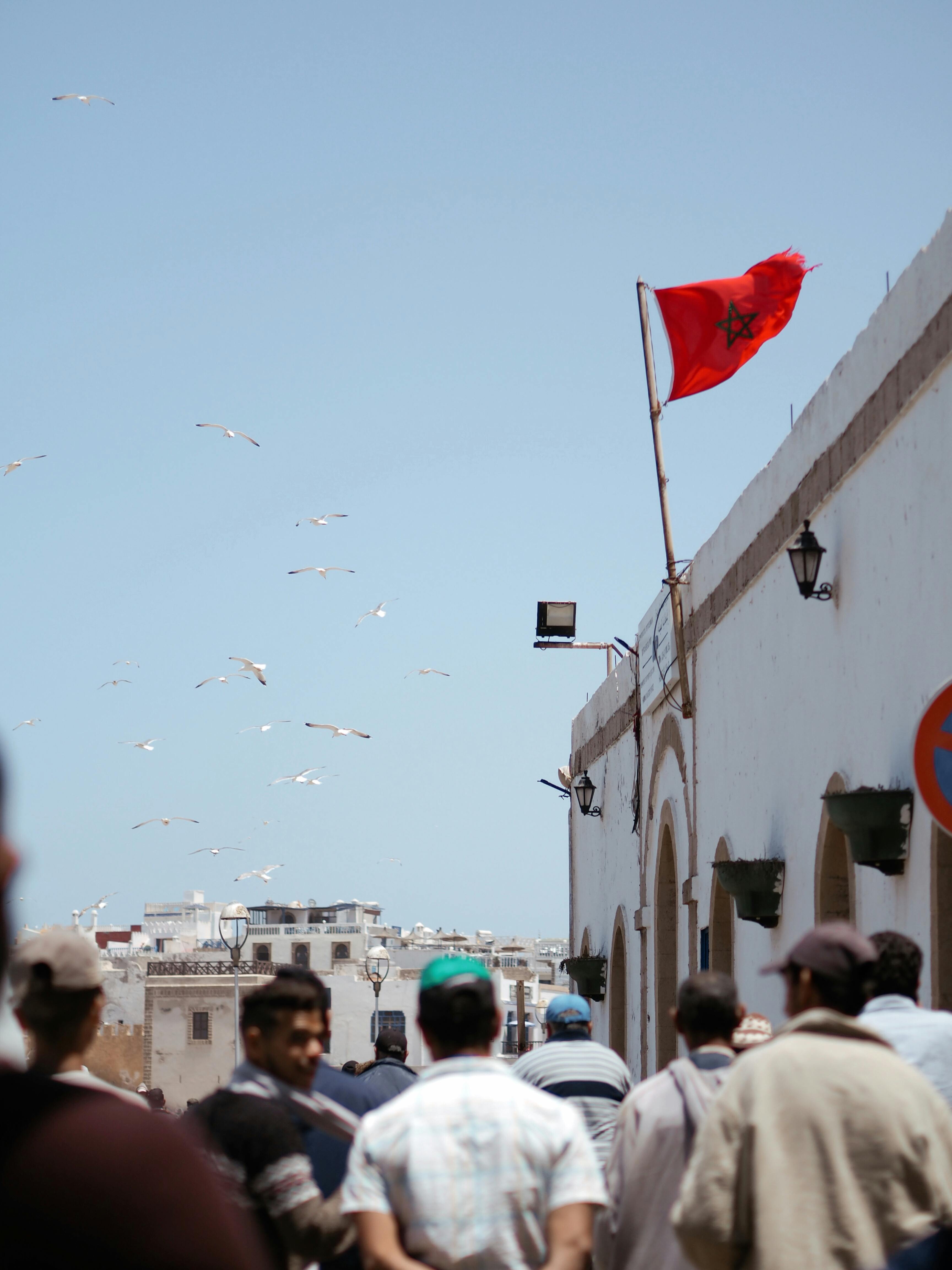 A bustling urban street scene with a Moroccan flag and birds in flight under a clear sky.