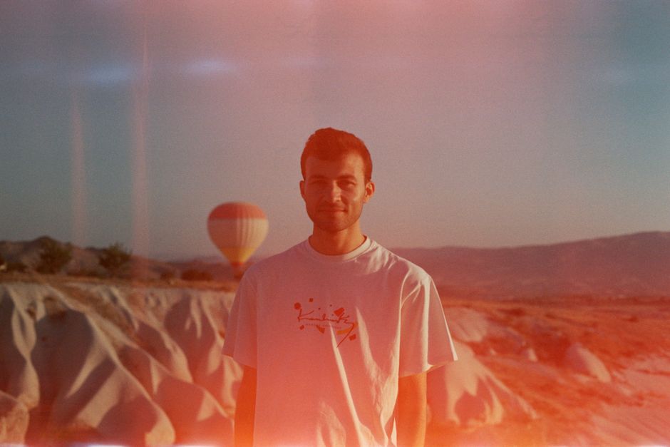 A man in a white t-shirt stands in a desert landscape with a hot air balloon in the background under a red filter effect.