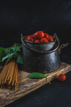 A rustic food arrangement featuring cherry tomatoes in a pot with spaghetti and basil leaves on a wooden board.